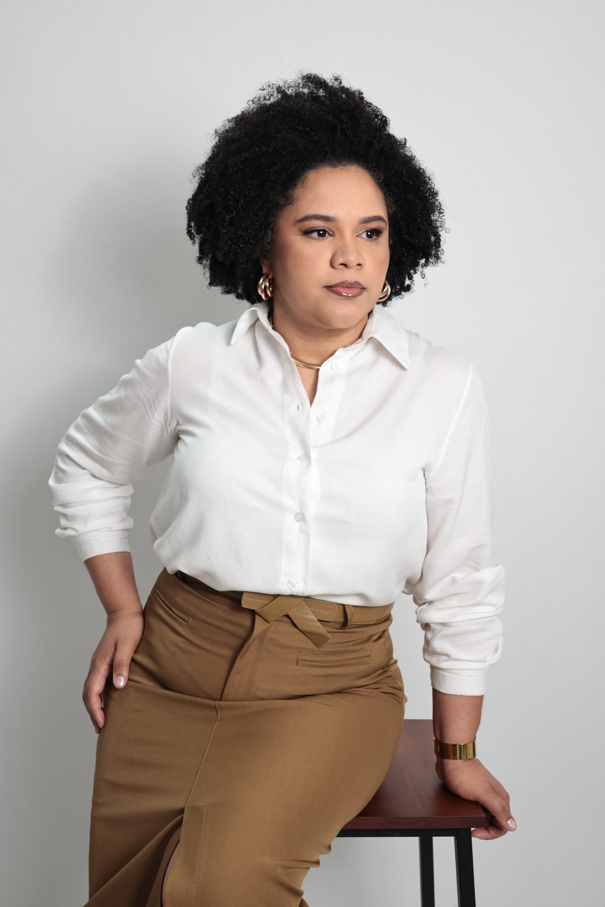 Retrato de mulher de camisa branca e calça caramelo sentada em banco, em fundo claro
