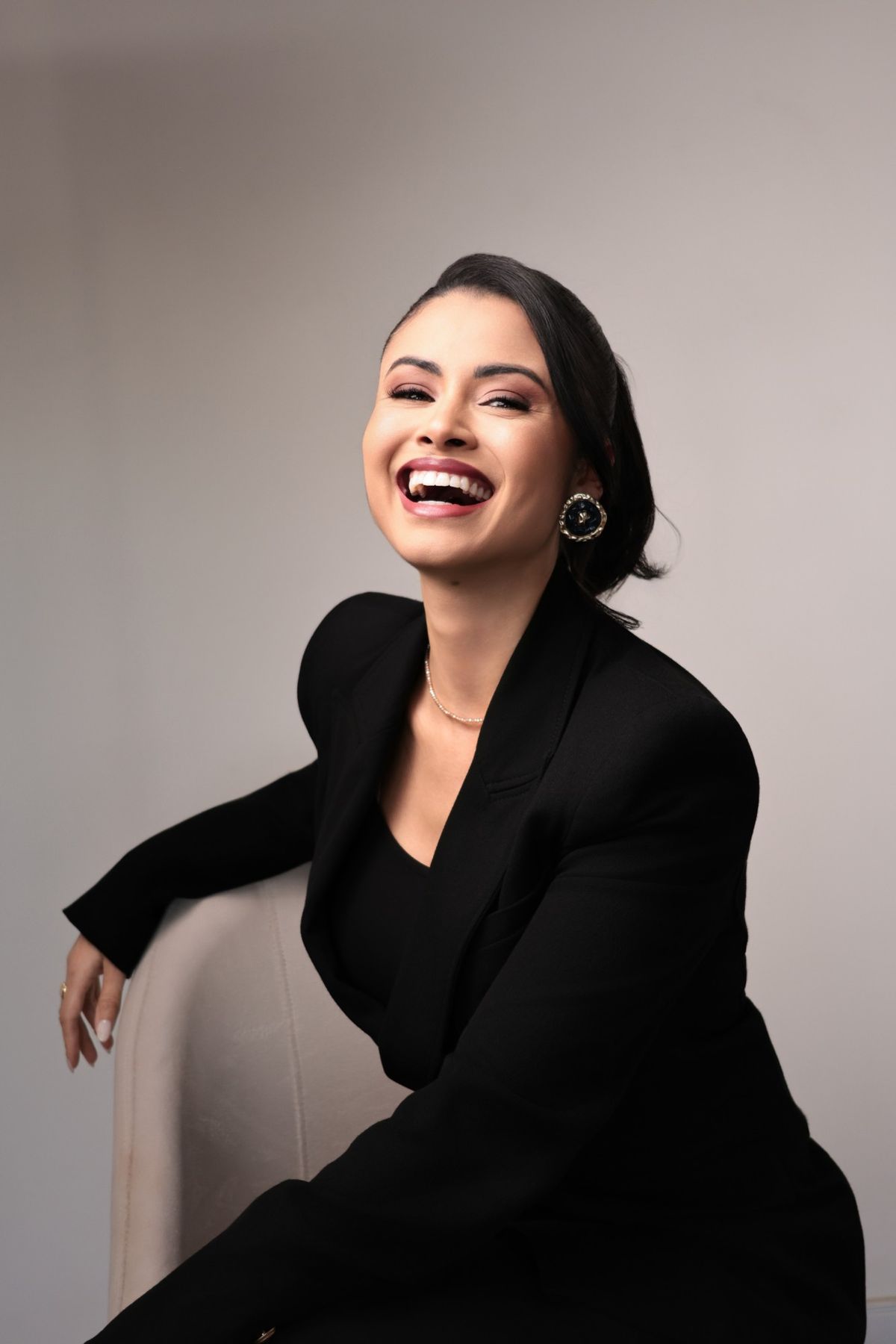 Retrato de mulher rindo, de blazer preto, sentada em poltrona clara